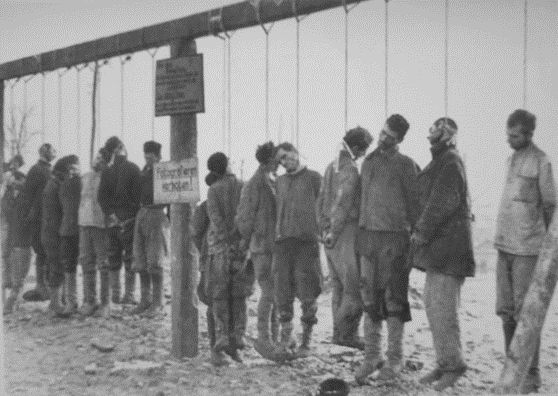 Os corpos de guerrilheiros soviéticos enforcados e uma placa dizendo "Não tire fotos!" (“Fotografieren proibido!”). Outra placa contém um texto em alemão e uma tradução em russo: “Somos bandidos, matamos e roubamos não apenas soldados alemães, mas também cidadãos russos”. https://xn--80aabgieomn8afgsnjq.xn--p1ai/fotografii-poveshennyh-kak-trofei-naczistskie-kazni-sovetskih-zhitelej/